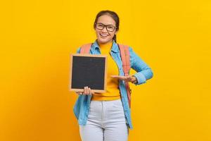 portret van lachende jonge Aziatische vrouw student in casual kleding met rugzak wijzend op leeg schoolbord met palm geïsoleerd op gele achtergrond. onderwijs in universiteitsconcept foto