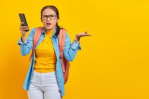 portret van verwarde jonge aziatische vrouw student in casual kleding met rugzak met mobiele telefoon en nee zeggen met handpalmen geïsoleerd op gele achtergrond. onderwijs in universiteitsconcept foto