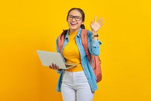 portret van vrolijke jonge Aziatische vrouw student in casual kleding met rugzak met behulp van laptop en gebaren zwaaiende hand geïsoleerd op gele achtergrond. onderwijs in universiteitsconcept foto