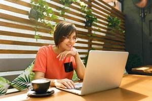 portret van zelfverzekerde volwassen professionele vrouw met een bril, een koraal t-shirt zittend op een zomerterras in café, laptopcomputer gebruikt voor werk, vrolijk lachend binnenshuis foto