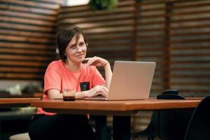 portret van zelfverzekerde volwassen professionele vrouw met een bril, een koraal t-shirt zittend op een zomerterras in café, laptopcomputer gebruikt voor werk, vrolijk lachend binnenshuis foto