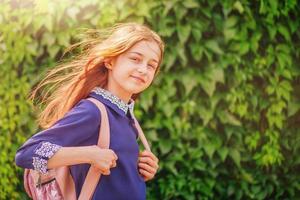 een schoolmeisje in een blauwe jurk met een witte kraag en een rugzak op een achtergrond van groen gebladerte. foto