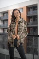 prachtige glamour brunette vrouw met geruite jas poseren op modern balkon met prachtig uitzicht op de stad. portret van een stijlvolle modieuze vrouw met lange benen, zwarte jeans dragen in balkon foto