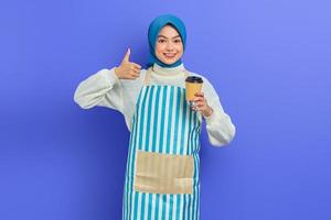 lachende jonge Aziatische moslimvrouw van in de twintig, gekleed in hijab en schort met papieren koffiekopje terwijl ze duim omhoog laat zien geïsoleerd over paarse achtergrond. mensen huisvrouw moslim lifestyle concept foto