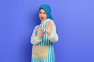 portret van boze jonge Aziatische moslimvrouw in hijab en gestreepte schort met stopgebaar met palm weigeren nee zeggen geïsoleerd op paarse achtergrond. mensen huisvrouw moslim lifestyle concept foto