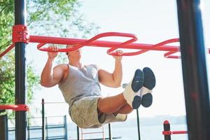 gespierde man met mooie torso oefenen op horizontale balken op de achtergrond van een wazig park. jonge man doet pull-ups buitenshuis foto