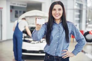 een mooie vrouw houdt een visitekaartje van het autoservicecentrum. de monteur inspecteert de auto onder de motorkap op de achtergrond