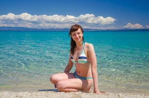 jong mooi meisje in bikini zit op zandstrand in de buurt van turquoise water van toroneos kolpos golf foto