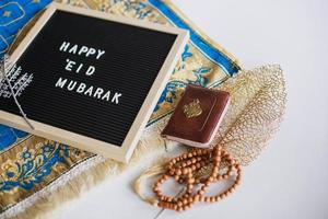 letterbord zegt happy eid mubarak op de gebedsmat met heilige boek al koran en gebedskralen. er is een Arabische letter die het heilige boek betekent foto