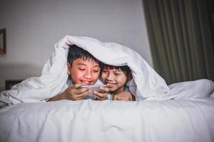 gelukkige Aziatische broer die online game speelt op een smartphone op het bed en hun lichaam bedekt met een deken foto