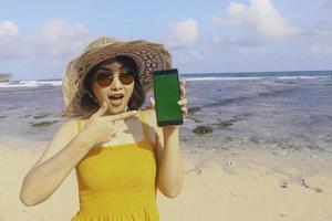 portret van een gelukkige glimlachende aziatische vrouw op het strand met een wijzend groen scherm van een smartphone foto