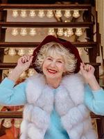 senior stijlvolle vrouw in bontjas en met grijze haren zittend op carrousel met paarse hoed. reizen, plezier, mode, geluk, seizoensconcept foto