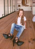 grappig klein meisje in wit t-shirt en spijkerbroek zittend op de vloer in hoge hakken schoenen in kledingkast dromen om volwassen en modieus te zijn. leeftijd, jeugd, mode, stijl, liefde hakken schoenen concept foto