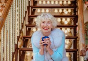 senior stijlvolle vrouw in bontjas en met grijze haren zittend op carrousel thee drinken en genieten van het leven. reizen, plezier, geluk, seizoensgebonden concept foto