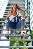 zwarte vrouw met gekleurde vlechten, haar smartphone raadplegend met haar voeten op een skateboard. foto