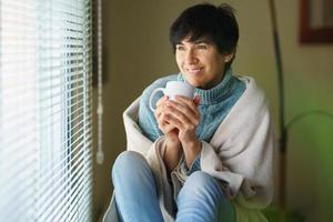 gelukkige vrouw van middelbare leeftijd die lacht bij een raam en een kopje koffie drinkt. foto