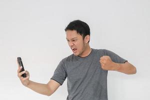 boze aziatische jongeman wordt boos op de smartphone. indonesië man dragen zwart shirt geïsoleerd grijze achtergrond. foto