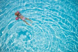 schattig klein peutermeisje dat in turquoise zwembad zwemt met opblaasbare armen aids-ondersteuning, bovenaanzicht. foto