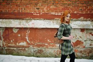 een openluchtportret van een jong mooi meisje met rood haar die geruite kleding met girly rugzakken dragen die zich op de bakstenen muurachtergrond in de winterdag bevinden. foto