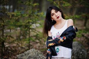 brunette zigeunermeisje gesteld tegen stenen op park. model slijtage op leren jas en t-shirt met ornament, broek. foto