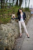 brunette zigeunermeisje met yorkshire terrier hond gesteld tegen stenen op park. model slijtage op leren jas en t-shirt met ornament, broek en schoenen met hoge hakken. foto