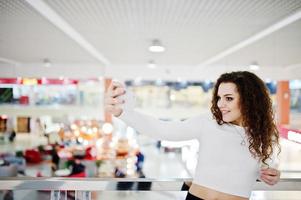 jong krullend model meisje selfie maken bij groot winkelcentrum in de buurt van glazen balustrades. foto