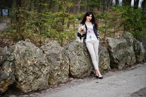 brunette zigeunermeisje met yorkshire terrier hond gesteld tegen stenen op park. model slijtage op leren jas en t-shirt met ornament, broek en schoenen met hoge hakken. foto