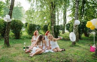 vijf meisjes droegen op witte jurk op vrijgezellenfeest buiten zitten en selfie maken in het park. foto