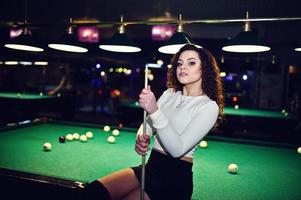 jong krullend meisje poseerde in de buurt van biljarttafel. sexy model bij zwarte mini minirok spelen russisch snooker. speel spel en leuk concept. foto