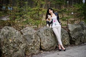 brunette zigeunermeisje met yorkshire terrier hond gesteld tegen stenen op park. model slijtage op leren jas en t-shirt met ornament, broek en schoenen met hoge hakken. foto