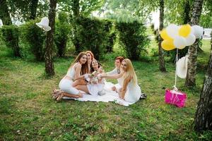 vijf meisjes met champagne bij de hand droegen op witte jurk op vrijgezellenfeest buiten in het park zitten en plezier hebben. foto