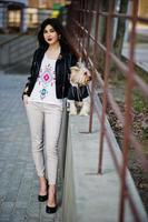 brunette zigeunermeisje met yorkshire terrier hond gesteld tegen stalen balustrades. model slijtage op leren jas en t-shirt met ornament, broek en schoenen met hoge hakken. foto