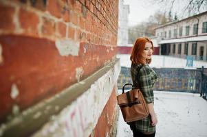 een openluchtportret van een jong mooi meisje met rood haar die geruite kleding met girly rugzakken dragen die zich op de bakstenen muurachtergrond in de winterdag bevinden. foto