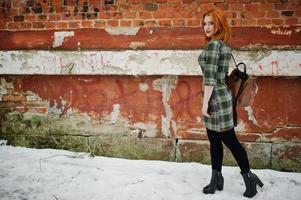 een openluchtportret van een jong mooi meisje met rood haar die geruite kleding met girly rugzakken dragen die zich op de bakstenen muurachtergrond in de winterdag bevinden. foto