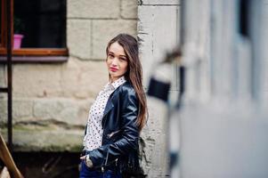 portret van stijlvol jong meisje draagt een leren jas en gescheurde jeans in de straten van de stad. straat fashion model stijl. foto