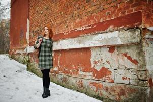 een openluchtportret van een jong mooi meisje met rood haar die geruite kleding met girly rugzakken dragen die zich op de bakstenen muurachtergrond in de winterdag bevinden. foto