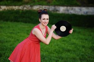 portret van jonge pinup meisje dragen bij retro vintage ouderwetse jurk in erwten met vinyl audio record, retro styling. foto