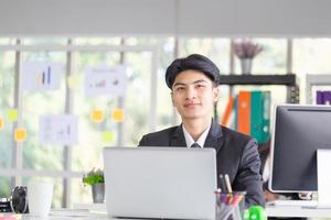 portret van een glimlachende jonge man die aan zijn bureau op kantoor zit, zakenman in pak die met een laptop op kantoor werkt foto