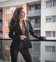 prachtige glamour brunette vrouw met zwarte jas poseren op modern balkon met prachtig uitzicht op de stad .portrait van een stijlvolle modieuze vrouw met lange benen, zwarte spijkerbroek dragen in balkon foto