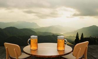 getapt of ambachtelijk bier in helder glas met bierschuim erop op een set houten tafels en stoelen. lounge zitplaatsen op het houten terras met bergachtergrond met godslicht of zonlicht.3D-rendering foto