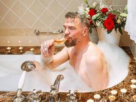 knappe volwassen man in de badkamer met schuim rose wijn drinken. spa, ontspannen, levensstijl, genieten van het levensconcept. foto