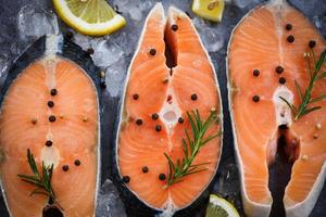 rauwe zalm visfilet en ijs voor gekookte steak zeevruchten - close-up verse zalm steak met kruiden en specerijen citroen rozemarijn op zwarte plaat achtergrond foto