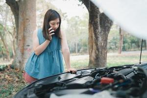 Aziatische vrouw die garage belt nadat de auto kapot gaat. vrouw die de motorkap opent en de verzekering of iemand belt om te helpen nadat de auto kapot is, parkeer aan de kant van de weg. vervoersconcept. foto