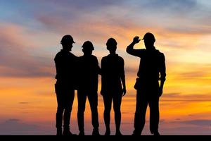 silhouet van ingenieur en arbeidersteam met het knippen van weg die zich met handen op schouders bevinden zonsondergangachtergrond, succes en groepswerkconcepten foto