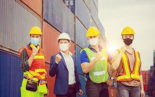 succes en teamwerkconcept, ingenieur en arbeidersteam die beschermingsgezichtsmasker dragen tegen coronavirus, gelukkige zakenmensenteamviering met vage vrachtcontainersachtergrond foto