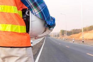 close-up van bouwvakker, ingenieur, werknemer die reddingsvesten draagt en witte veiligheidshelmen vasthoudt voor veiligheid op het werk, staande aan de kant van de weg.