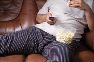 een luie man die de afstandsbediening van de tv vasthoudt en popcorn eet foto