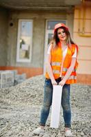 ingenieur bouwer vrouw in uniform vest en oranje beschermende helm houdt zakelijke tekenpapierrol vast tegen nieuwbouw. eigendom woonblok thema. foto