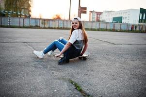 jonge stedelijke tienermeisje met skateboard, draag een bril, pet en gescheurde spijkerbroek op het sportveld van de werf bij zonsondergang. foto