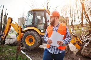 brute baard werknemer man pak bouwvakker in veiligheid oranje helm tegen traktor met plan papier bij de hand. foto